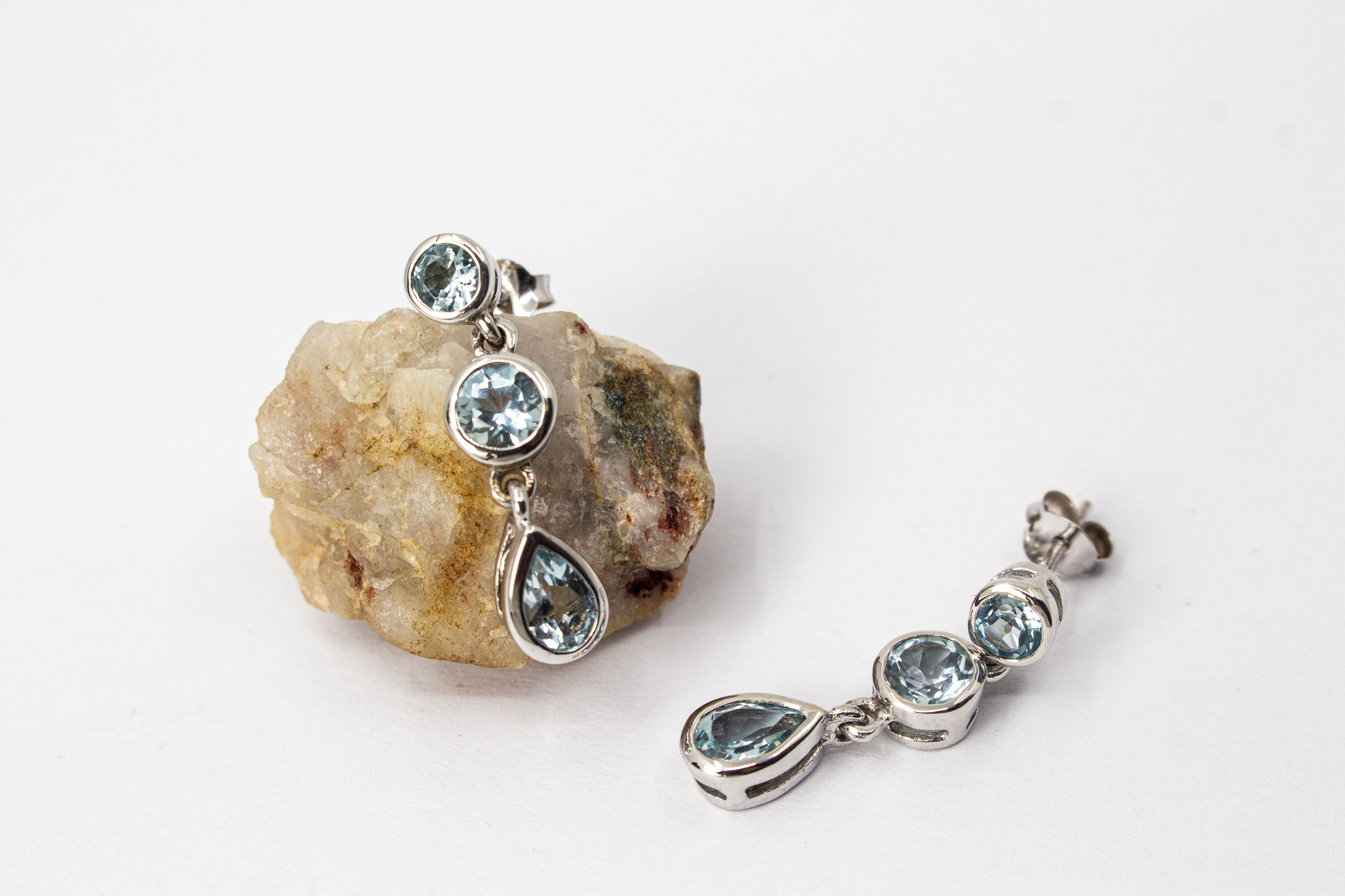 Ein Paar farbenfrohe Ohrstecker aus rhodiniertem 925er Sterlingsilber von der Goldschmiede Regina Kraus, präsentiert an einem transparenten Schmuckständer. Jeder Ohrhänger zeigt drei vertikal angeordnete, facettierte Edelsteine in Zargenfassung: ein hell