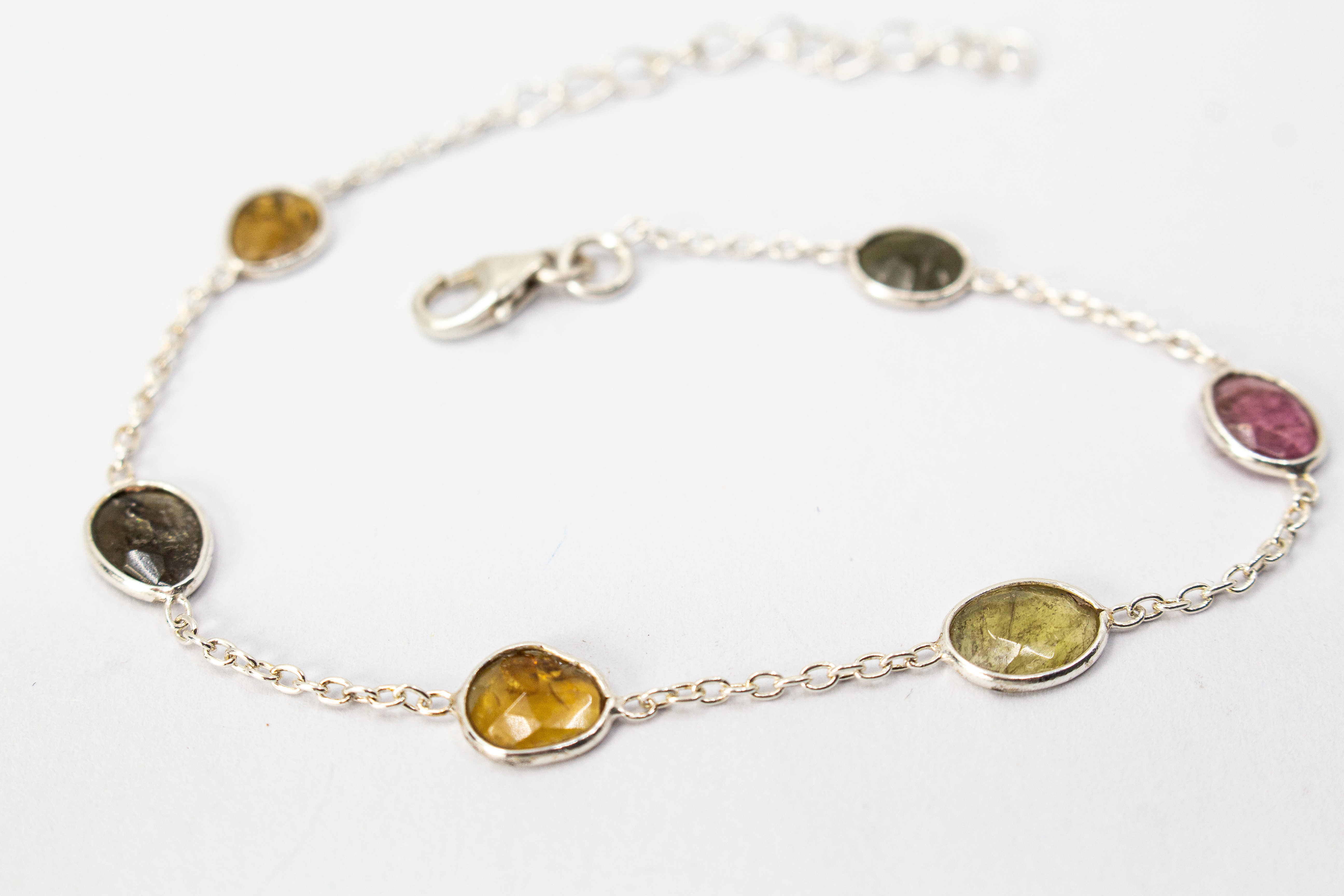 Ein filigranes Armband aus 925 Sterlingsilber von der Goldschmiede Regina Kraus, flach auf weißem Hintergrund arrangiert. In die feine Gliederkette sind sechs verschiedenfarbige, flache Turmaline in ovalen Zargenfassungen eingearbeitet – darunter Gelb-,