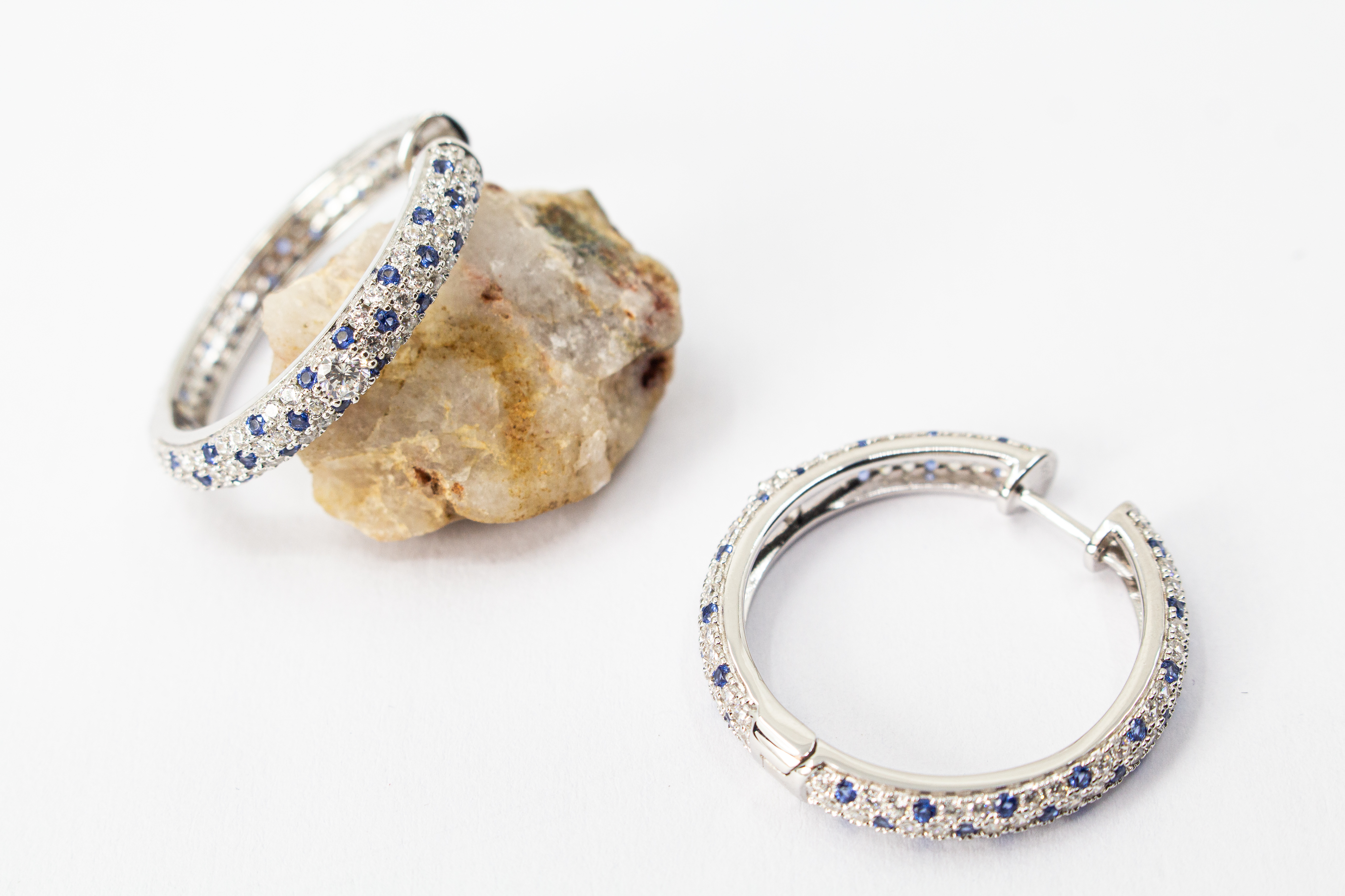 Ein Paar luxuriöse, rhodinierte Klappcreolen aus 925er Sterlingsilber von der Goldschmiede Regina Kraus. Die breite Außenseite ist dreireihig im edlen Pavé-Stil mit funkelnden, weißen und saphirblauen Zirkonia-Steinen besetzt. Eine Creole lehnt dekorativ
