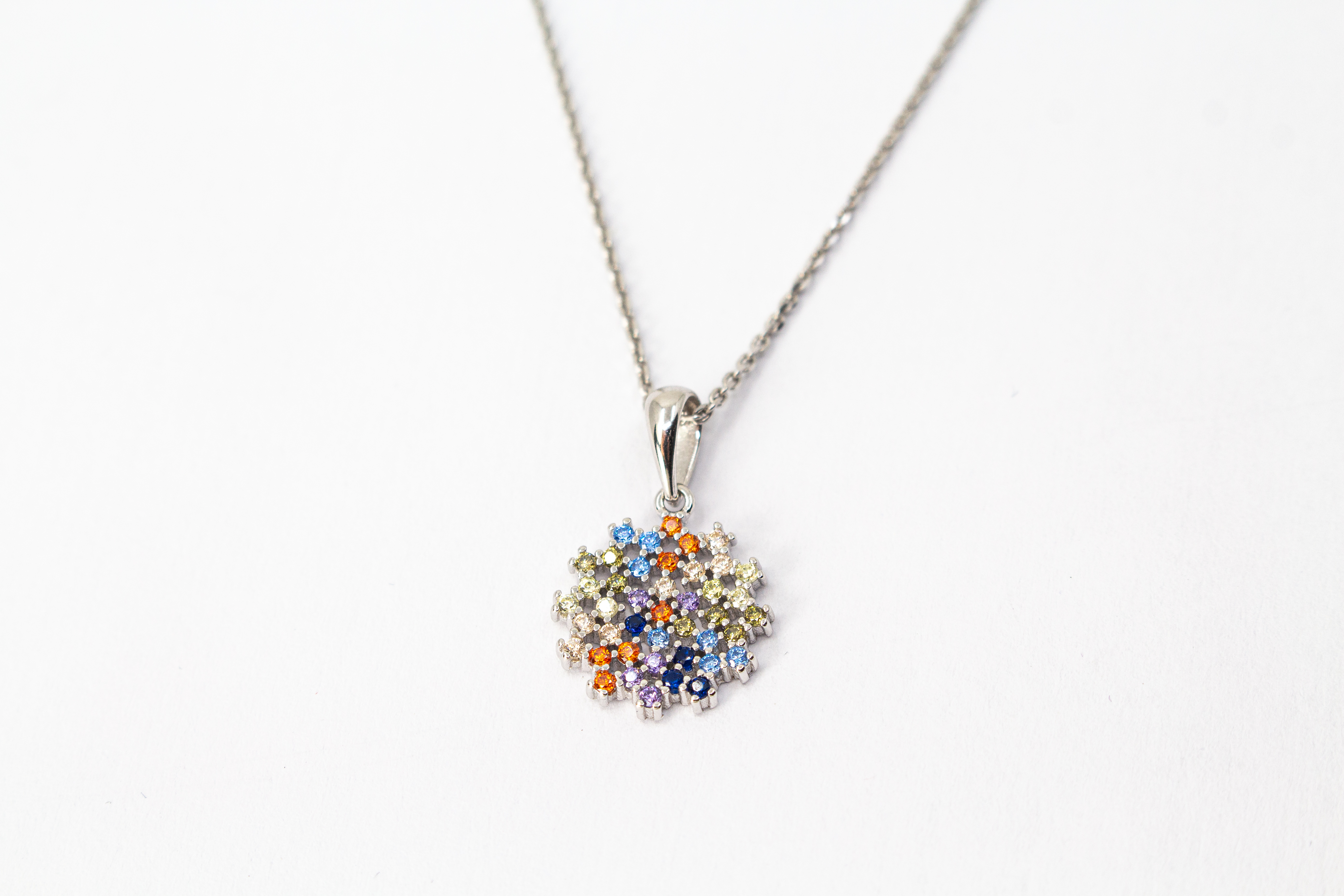 Eine feine Halskette aus rhodiniertem 925er Sterlingsilber von der Goldschmiede Regina Kraus, schräg auf weißem Hintergrund liegend. Der runde Anhänger ist filigran wie ein Mandala oder eine Blüte gestaltet und dicht mit kleinen, bunt funkelnden Zirkonia-