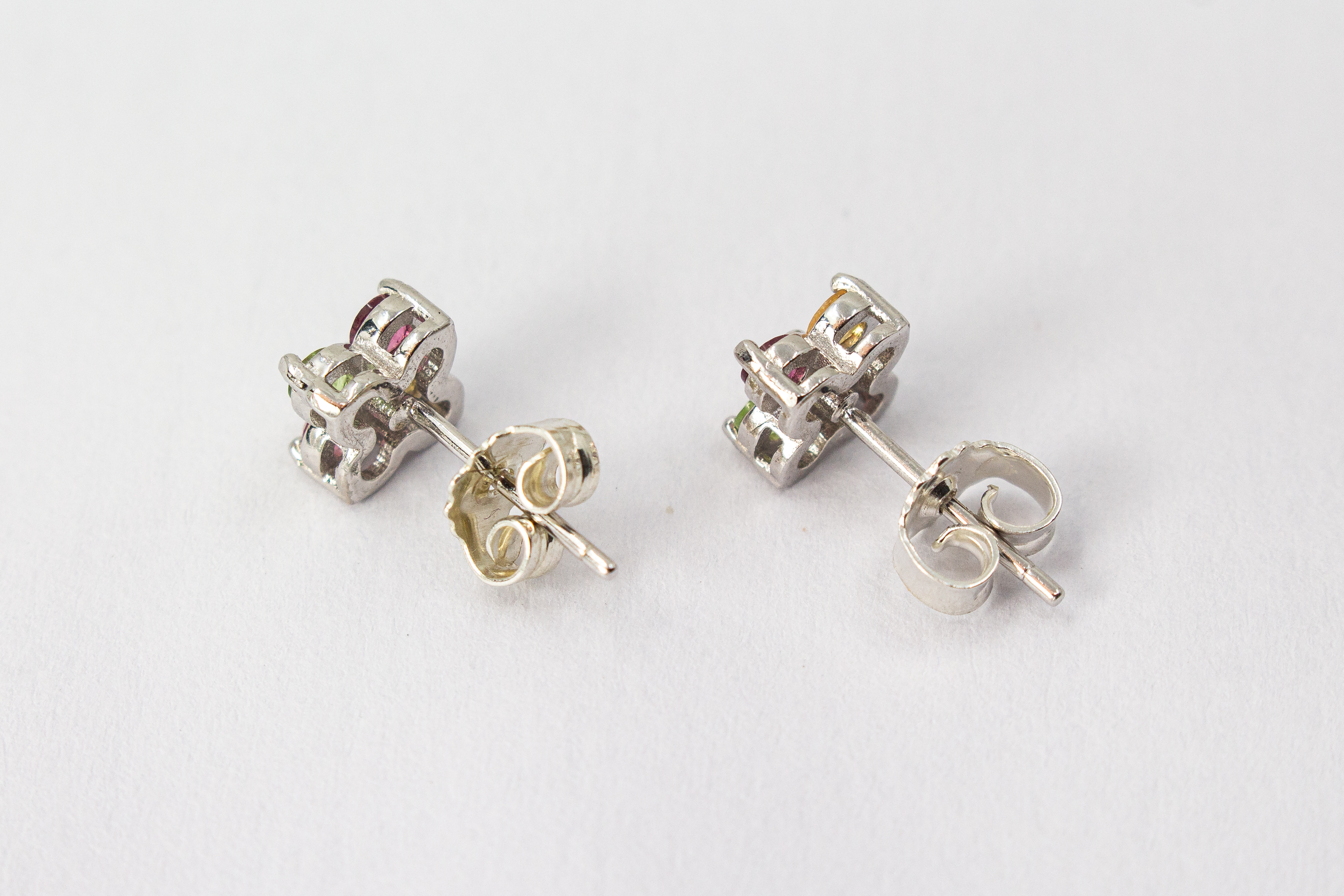 Die filigranen Silber-Ohrstecker „Elfenblüte“ aus der Goldschmiede Regina Kraus an einem transparenten Halter. Die Stecker aus rhodiniertem 925er Sterlingsilber zeigen eine zarte Blütenform, besetzt mit vier bunten Turmalinen in Rosé, Grün und Gelb um ei