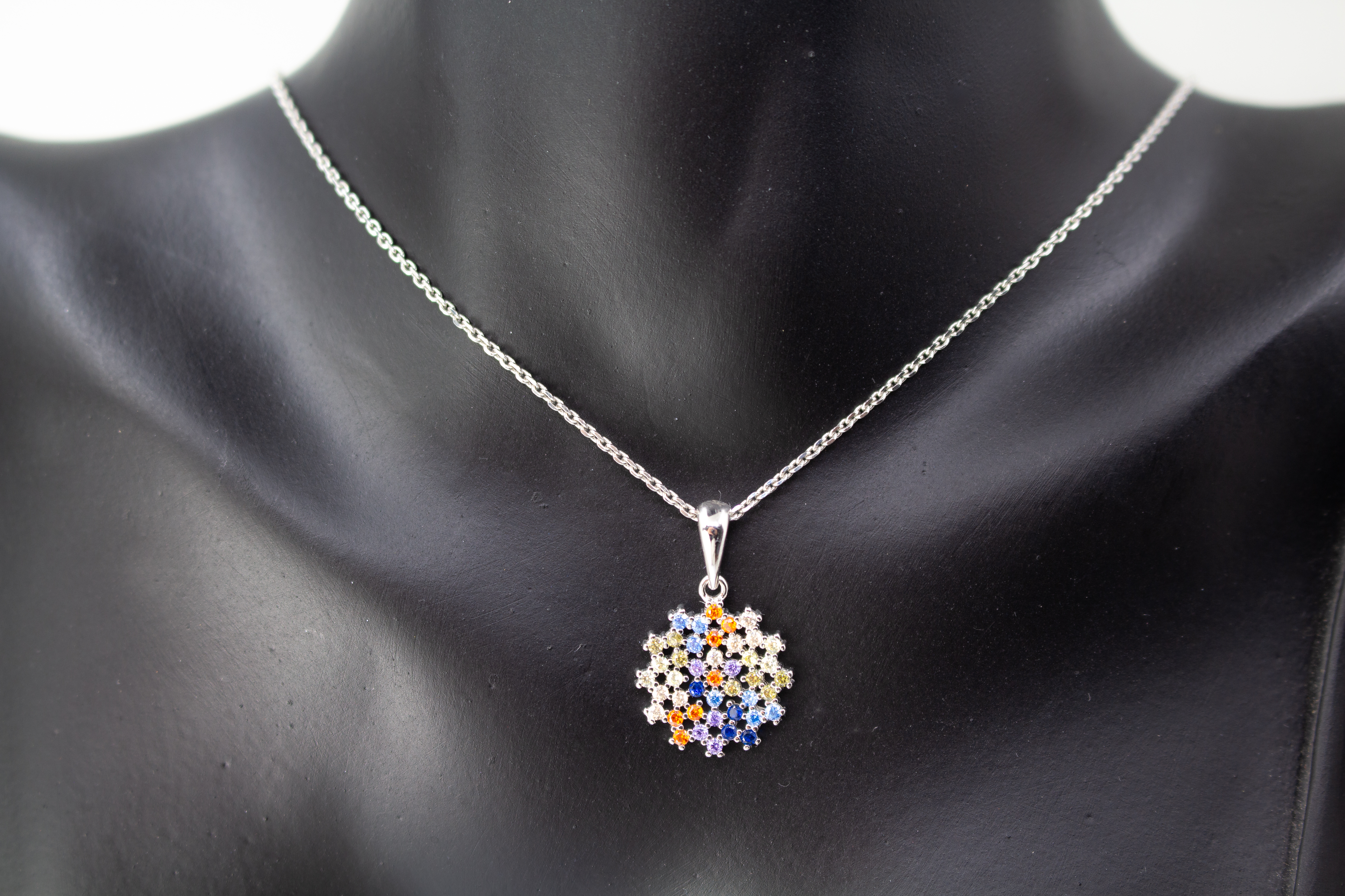 Eine feine Halskette aus rhodiniertem 925er Sterlingsilber von der Goldschmiede Regina Kraus, schräg auf weißem Hintergrund liegend. Der runde Anhänger ist filigran wie ein Mandala oder eine Blüte gestaltet und dicht mit kleinen, bunt funkelnden Zirkonia-