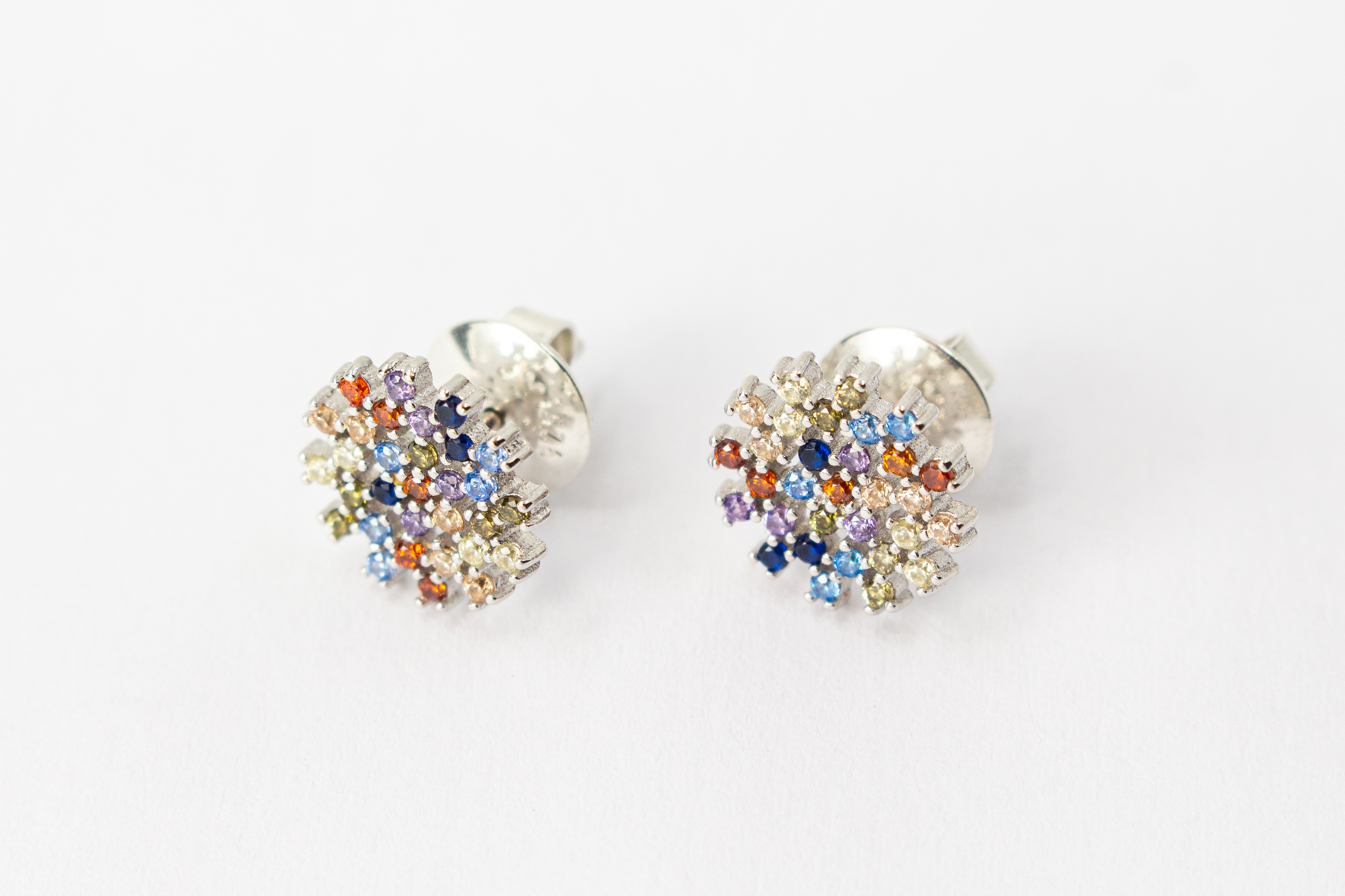 Die filigranen 925er Silber-Ohrstecker „Fleur de Lumière“ aus der Goldschmiede Regina Kraus, präsentiert auf einem transparenten Schmuckständer. Das florale Design erinnert an eine funkelnde Lichtblume und ist kunstvoll mit kleinen, bunt strahlenden Zirk