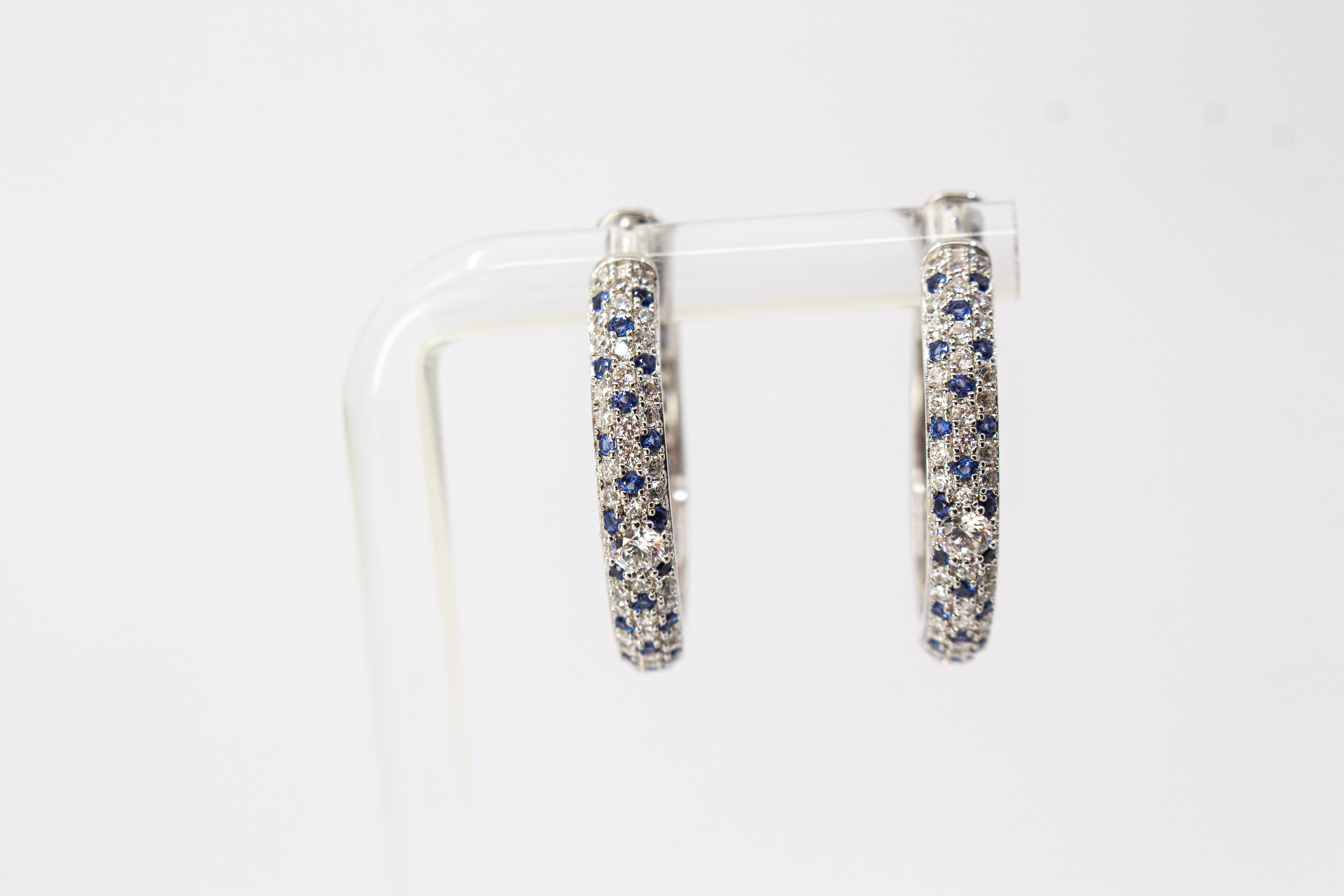 Ein Paar luxuriöse, rhodinierte Klappcreolen aus 925er Sterlingsilber von der Goldschmiede Regina Kraus. Die breite Außenseite ist dreireihig im edlen Pavé-Stil mit funkelnden, weißen und saphirblauen Zirkonia-Steinen besetzt. Eine Creole lehnt dekorativ