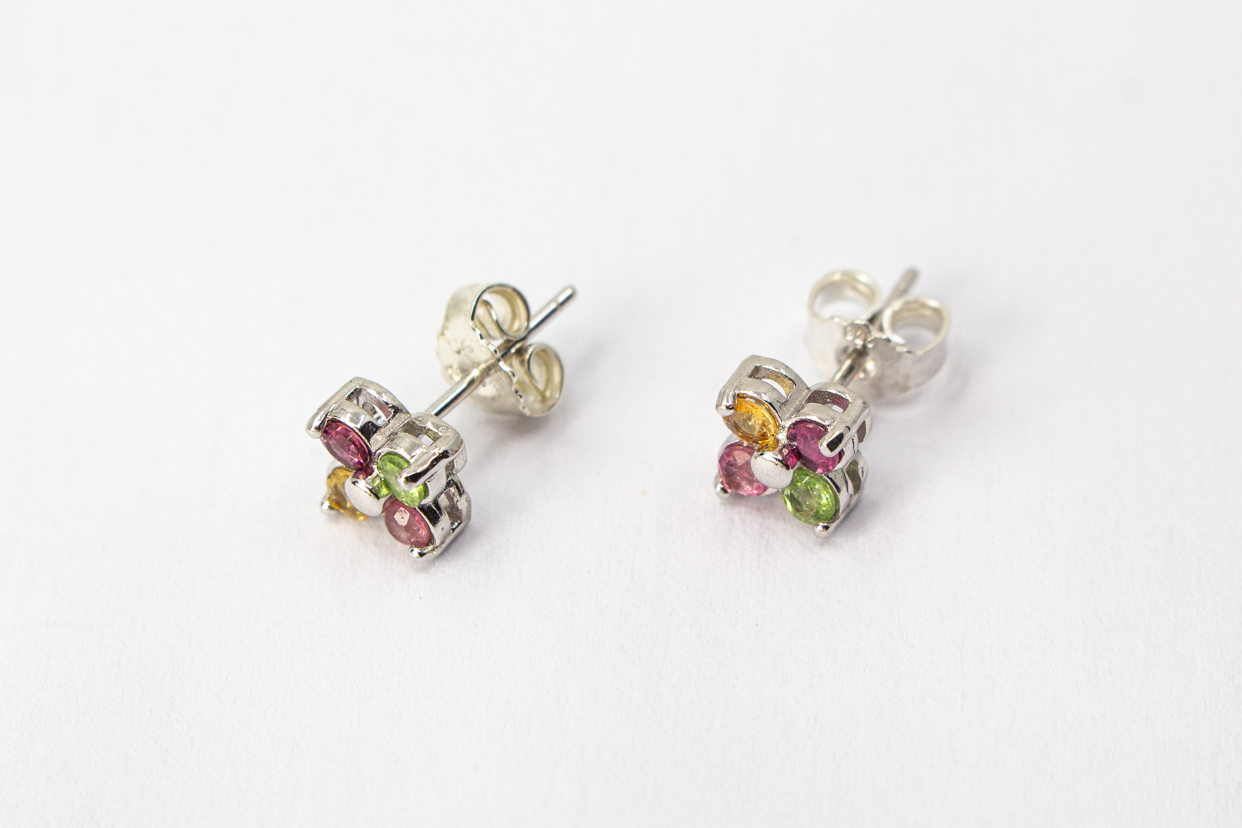 Die filigranen Silber-Ohrstecker „Elfenblüte“ aus der Goldschmiede Regina Kraus an einem transparenten Halter. Die Stecker aus rhodiniertem 925er Sterlingsilber zeigen eine zarte Blütenform, besetzt mit vier bunten Turmalinen in Rosé, Grün und Gelb um ei