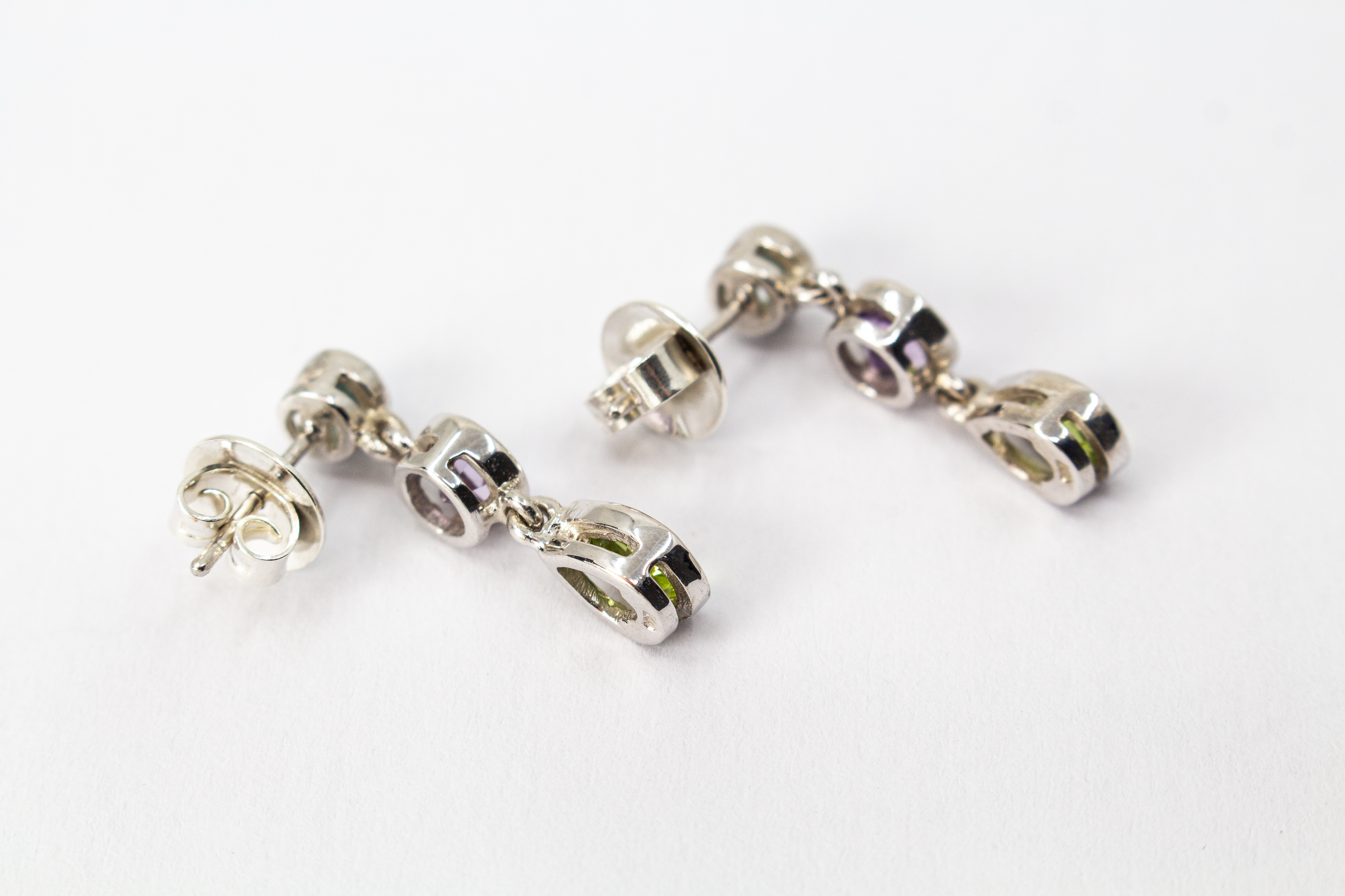 Ein Paar farbenfrohe Ohrstecker aus rhodiniertem 925er Sterlingsilber von der Goldschmiede Regina Kraus, präsentiert an einem transparenten Schmuckständer. Jeder Ohrhänger zeigt drei vertikal angeordnete, facettierte Edelsteine in Zargenfassung: ein hell