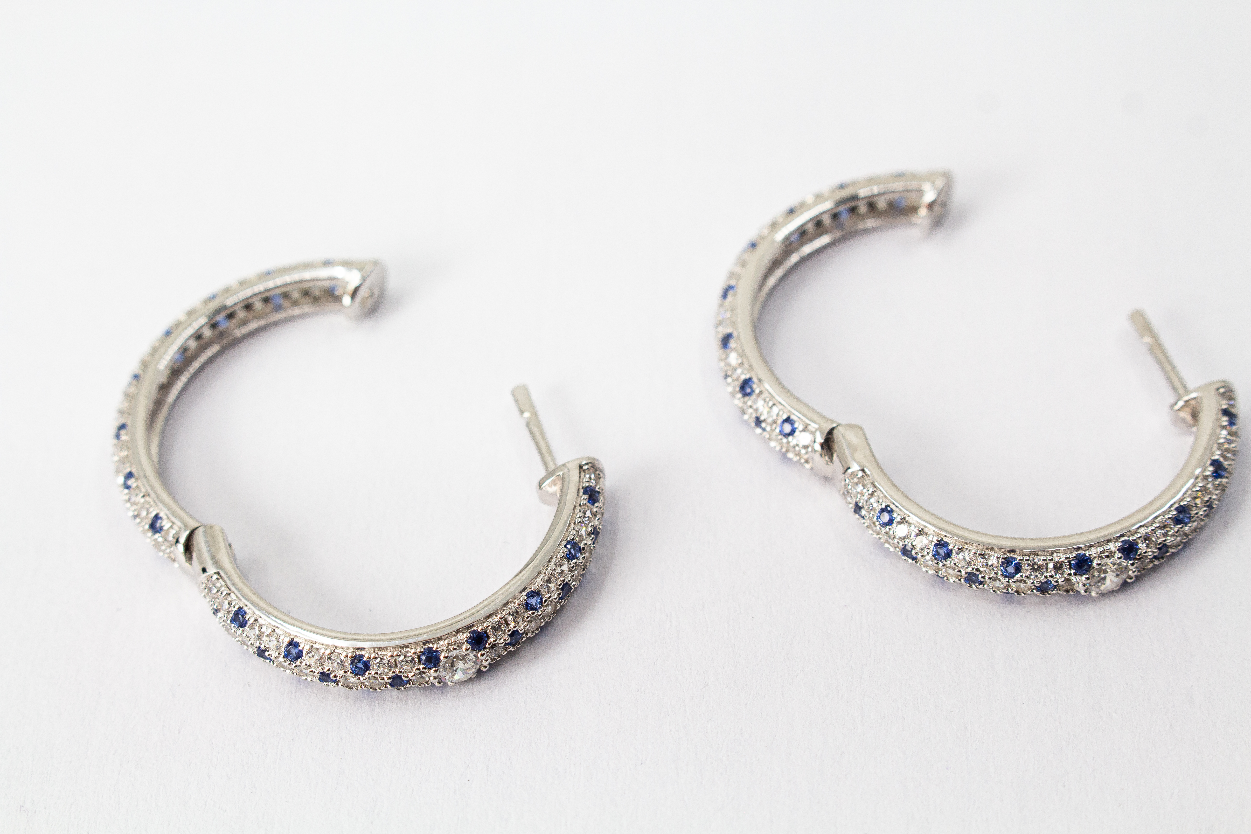 Ein Paar luxuriöse, rhodinierte Klappcreolen aus 925er Sterlingsilber von der Goldschmiede Regina Kraus. Die breite Außenseite ist dreireihig im edlen Pavé-Stil mit funkelnden, weißen und saphirblauen Zirkonia-Steinen besetzt. Eine Creole lehnt dekorativ