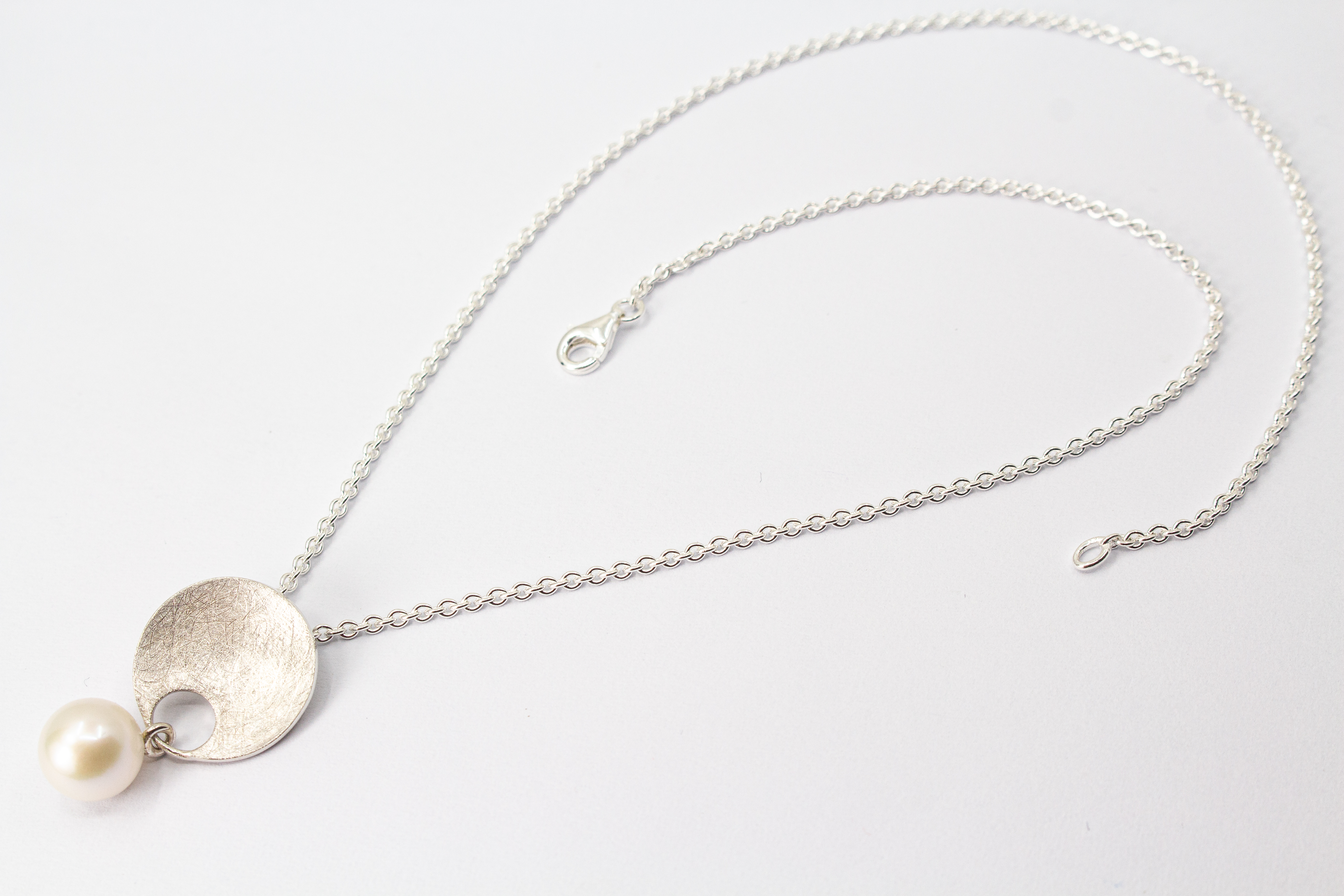 Feine Halskette aus 925 Sterlingsilber mit einem runden, matt strukturierten Scheibenanhänger, an dem eine weiße, runde Süßwasserzuchtperle beweglich befestigt ist, von der Goldschmiede Regina Kraus.