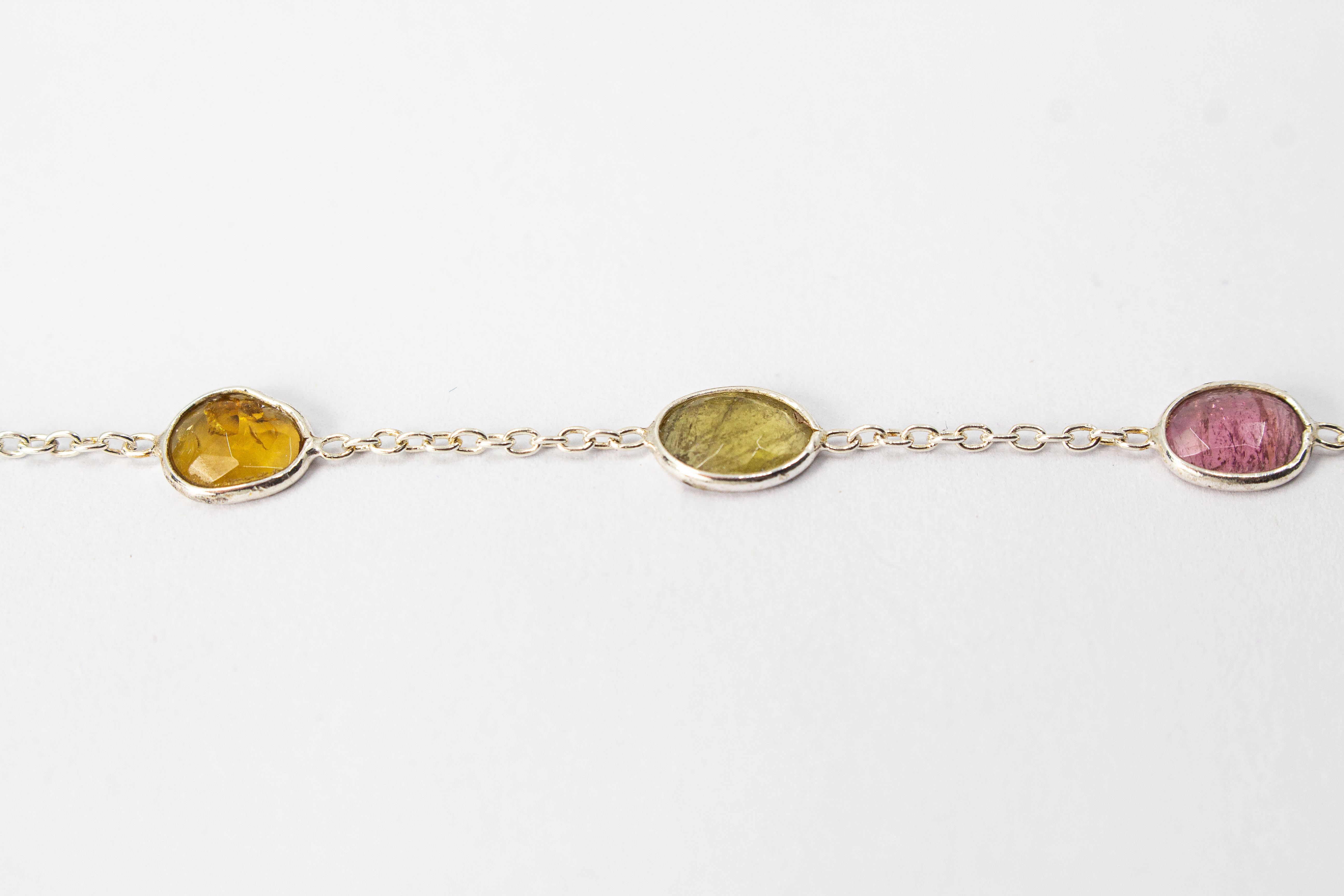 Ein filigranes Armband aus 925 Sterlingsilber von der Goldschmiede Regina Kraus, flach auf weißem Hintergrund arrangiert. In die feine Gliederkette sind sechs verschiedenfarbige, flache Turmaline in ovalen Zargenfassungen eingearbeitet – darunter Gelb-,