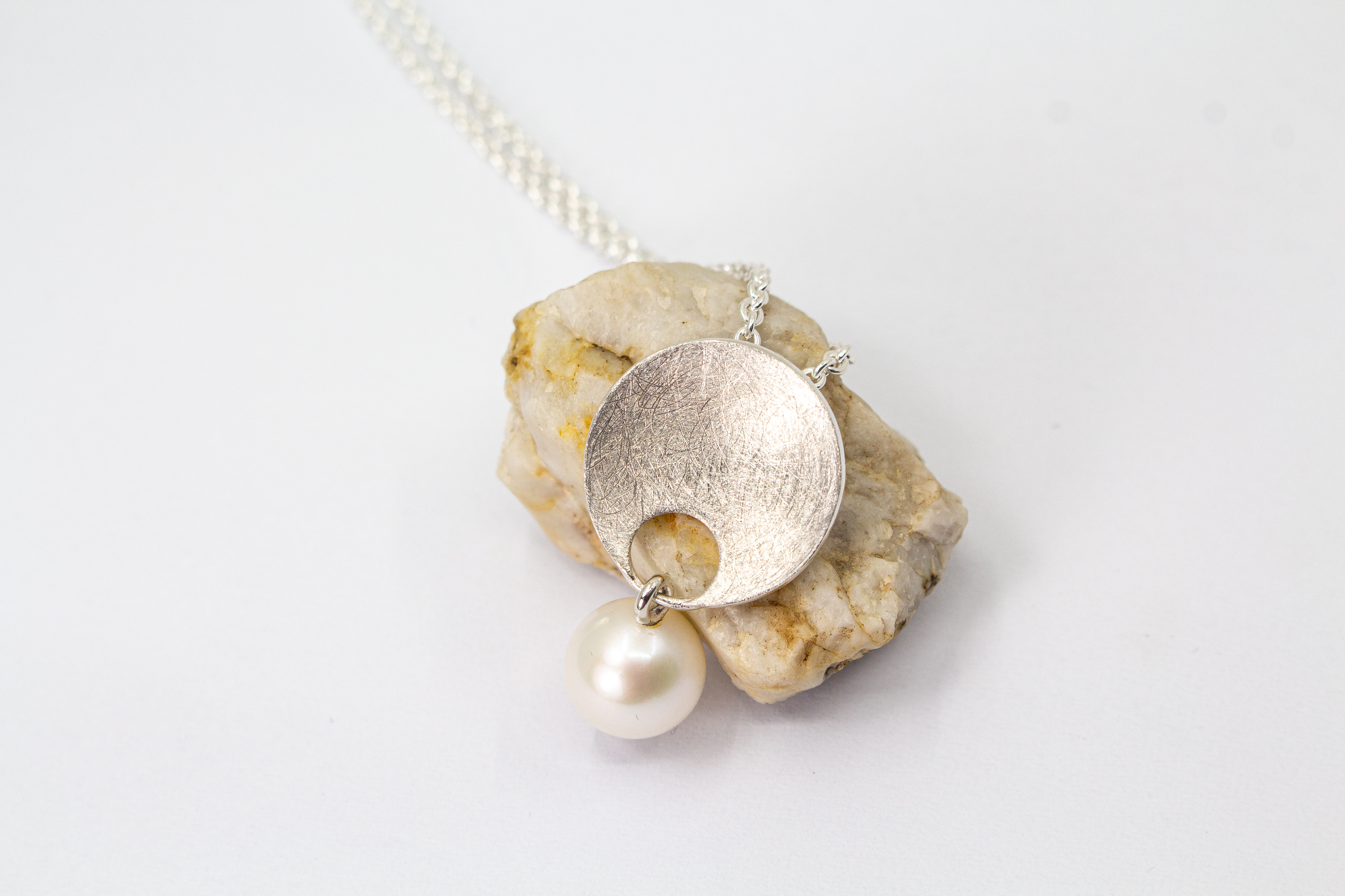Feine Halskette aus 925 Sterlingsilber mit einem runden, matt strukturierten Scheibenanhänger, an dem eine weiße, runde Süßwasserzuchtperle beweglich befestigt ist, von der Goldschmiede Regina Kraus.