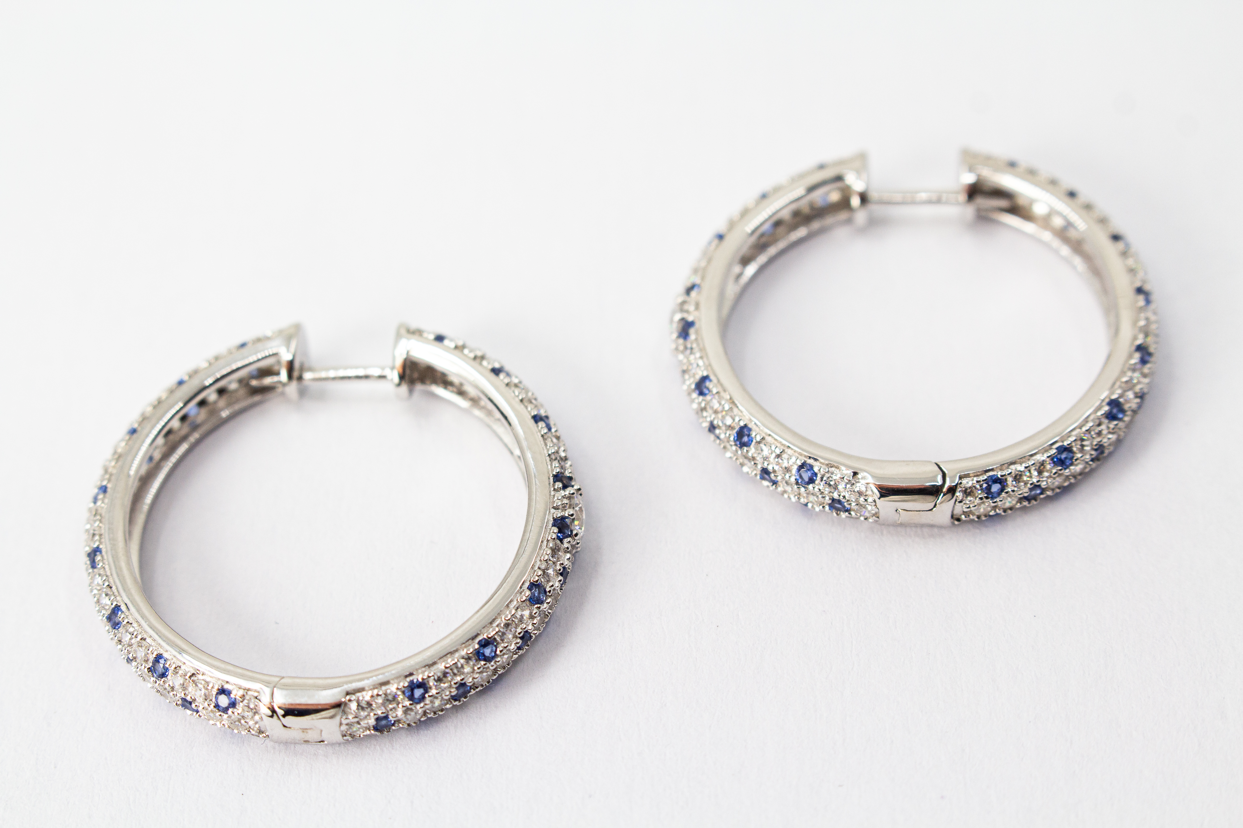 Ein Paar luxuriöse, rhodinierte Klappcreolen aus 925er Sterlingsilber von der Goldschmiede Regina Kraus. Die breite Außenseite ist dreireihig im edlen Pavé-Stil mit funkelnden, weißen und saphirblauen Zirkonia-Steinen besetzt. Eine Creole lehnt dekorativ