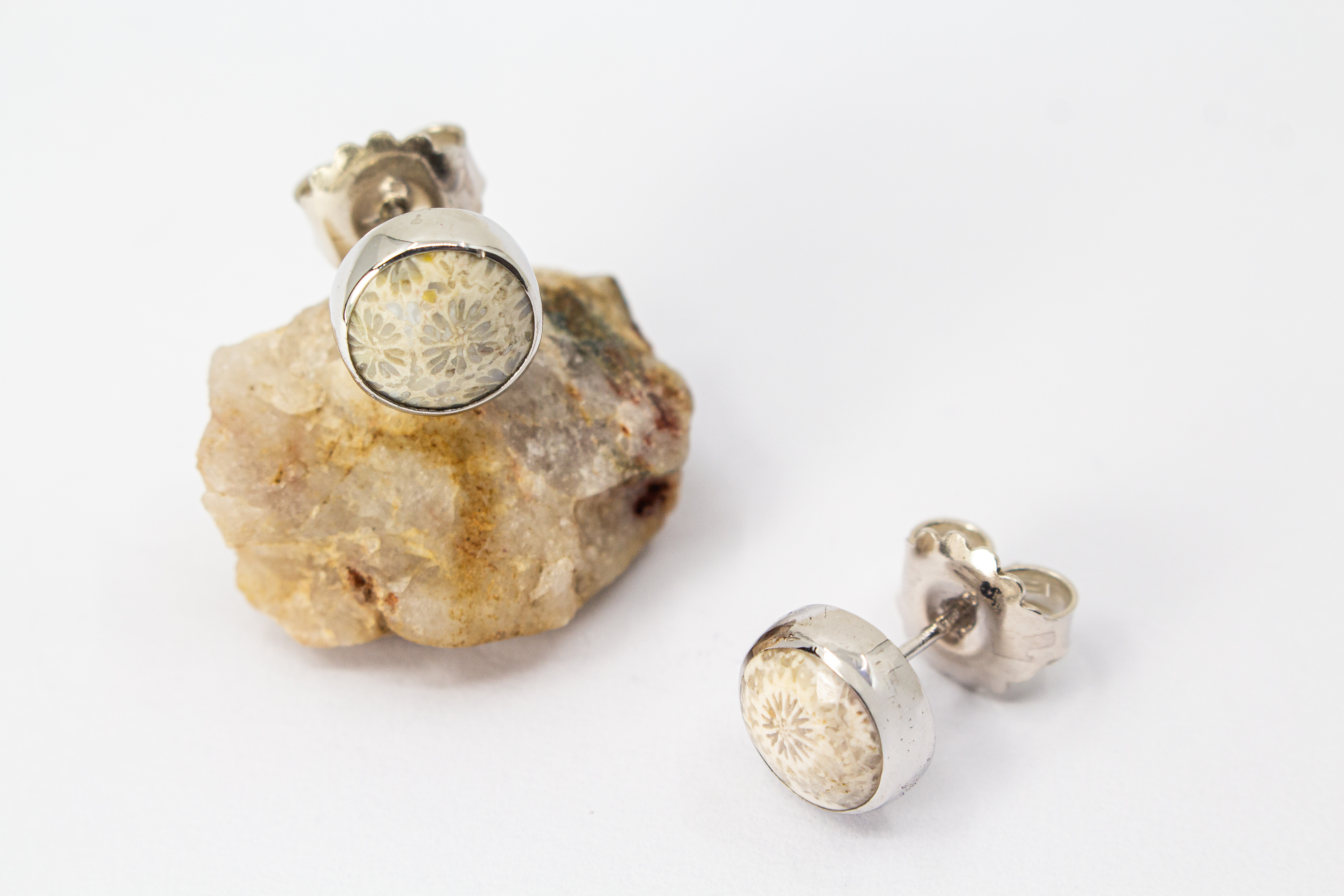 Ein Paar runde Damen-Ohrstecker aus poliertem 925er Sterling Silber, besetzt mit runden fossilen Korallen-Cabochons, die eine feine, beige-graue sternförmige Naturmusterung aufweisen, arrangiert neben einem hellen Rohstein