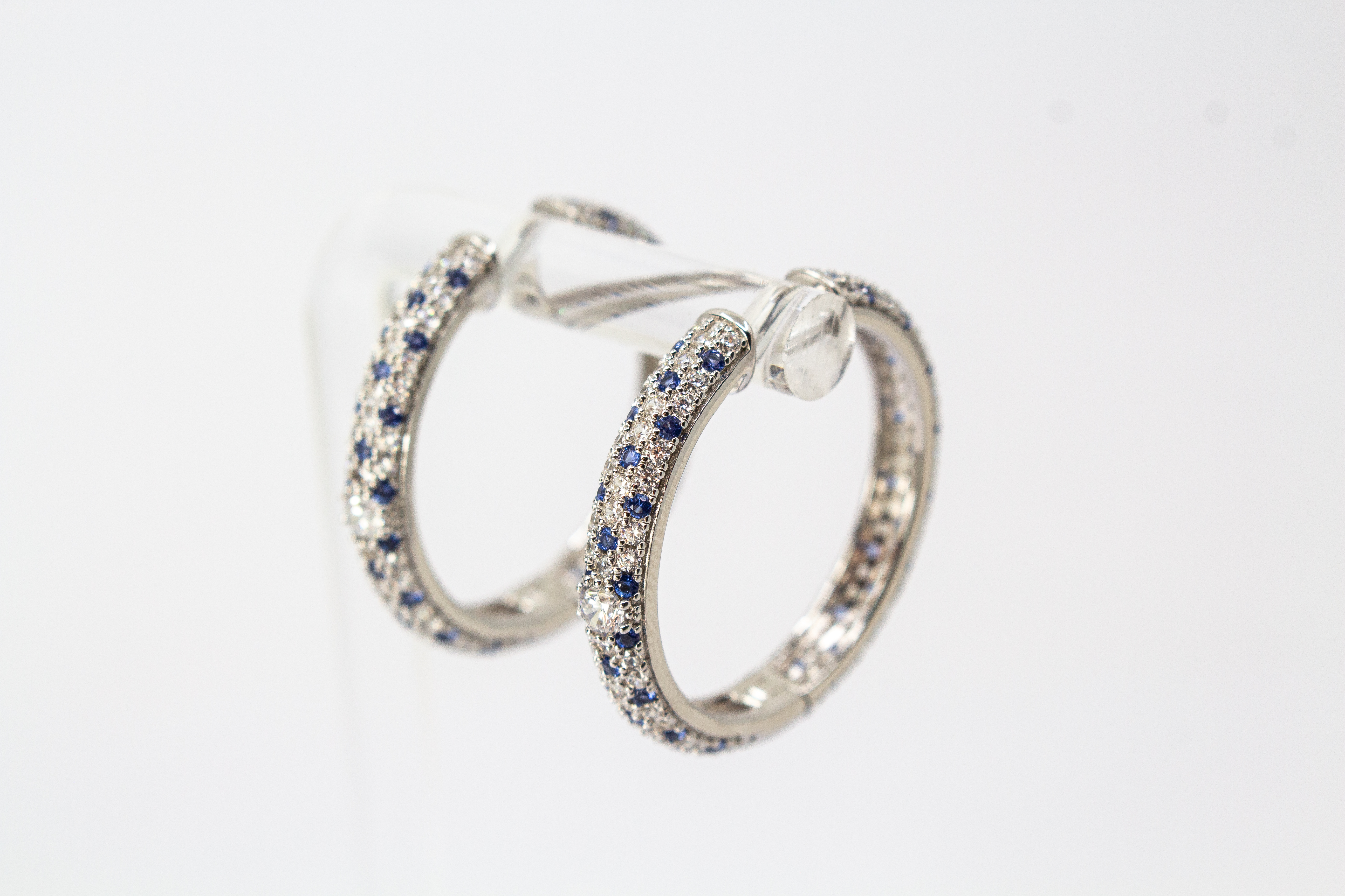 Ein Paar luxuriöse, rhodinierte Klappcreolen aus 925er Sterlingsilber von der Goldschmiede Regina Kraus. Die breite Außenseite ist dreireihig im edlen Pavé-Stil mit funkelnden, weißen und saphirblauen Zirkonia-Steinen besetzt. Eine Creole lehnt dekorativ