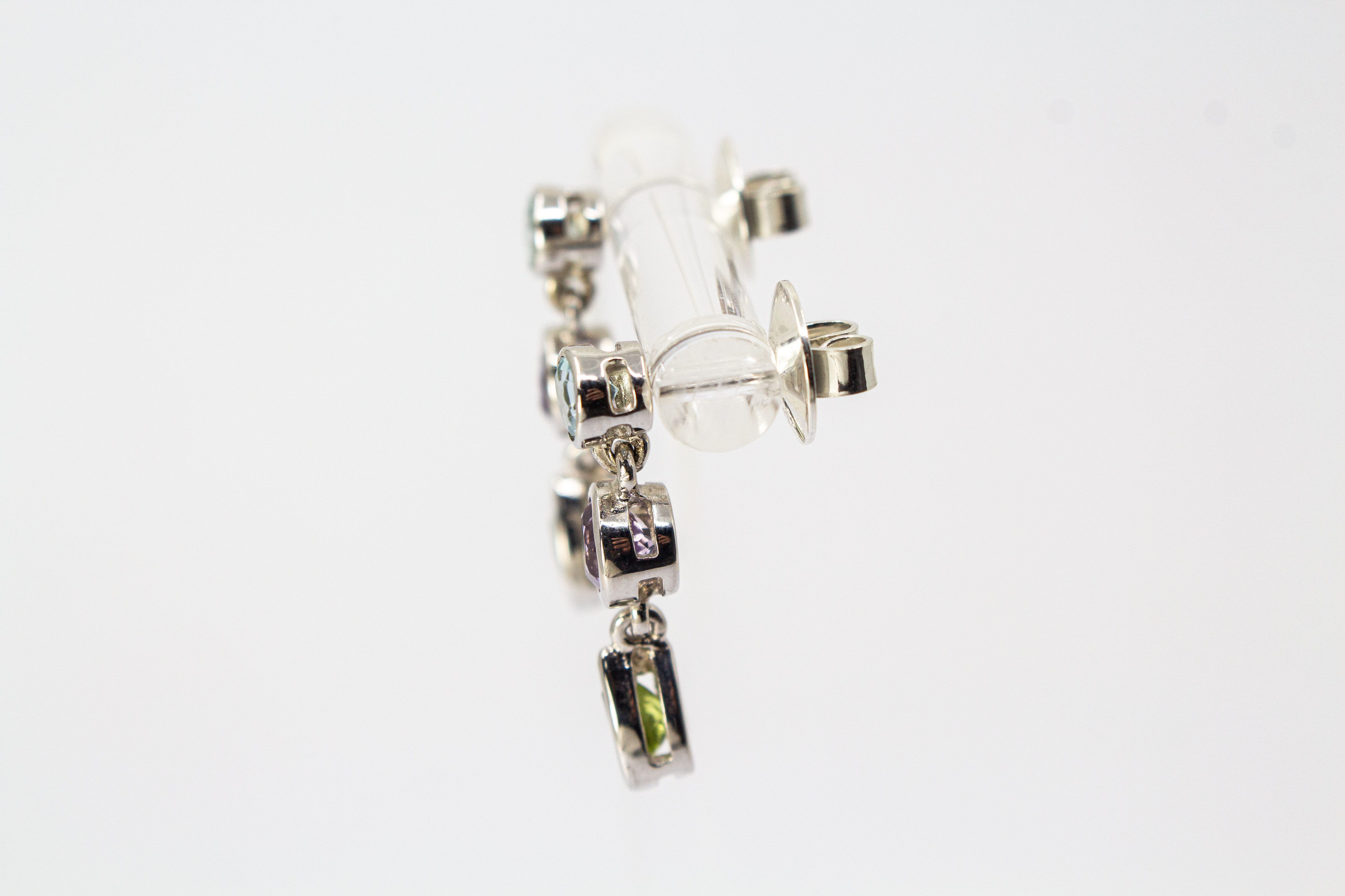 Ein Paar farbenfrohe Ohrstecker aus rhodiniertem 925er Sterlingsilber von der Goldschmiede Regina Kraus, präsentiert an einem transparenten Schmuckständer. Jeder Ohrhänger zeigt drei vertikal angeordnete, facettierte Edelsteine in Zargenfassung: ein hell