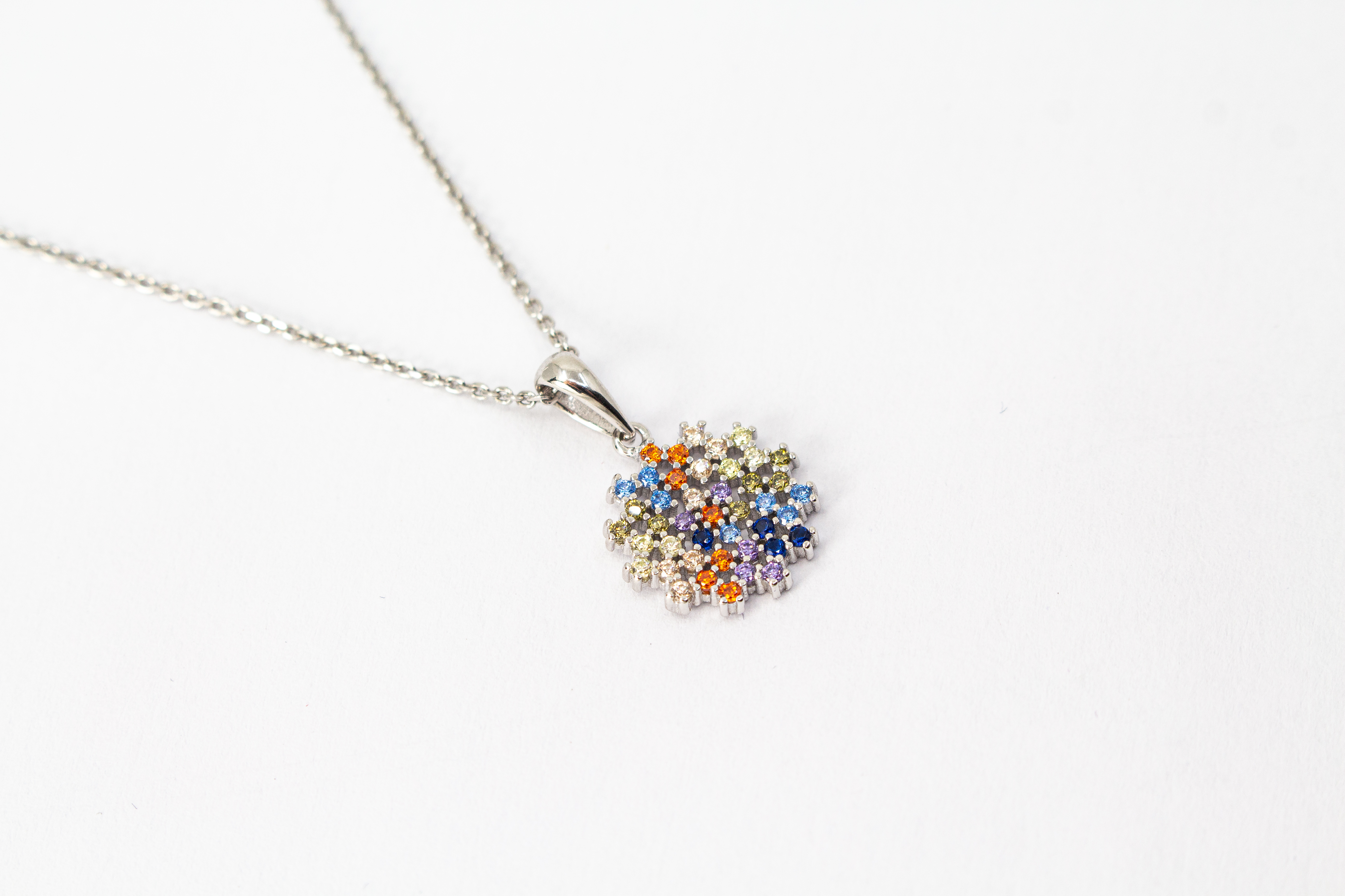 Eine feine Halskette aus rhodiniertem 925er Sterlingsilber von der Goldschmiede Regina Kraus, schräg auf weißem Hintergrund liegend. Der runde Anhänger ist filigran wie ein Mandala oder eine Blüte gestaltet und dicht mit kleinen, bunt funkelnden Zirkonia-