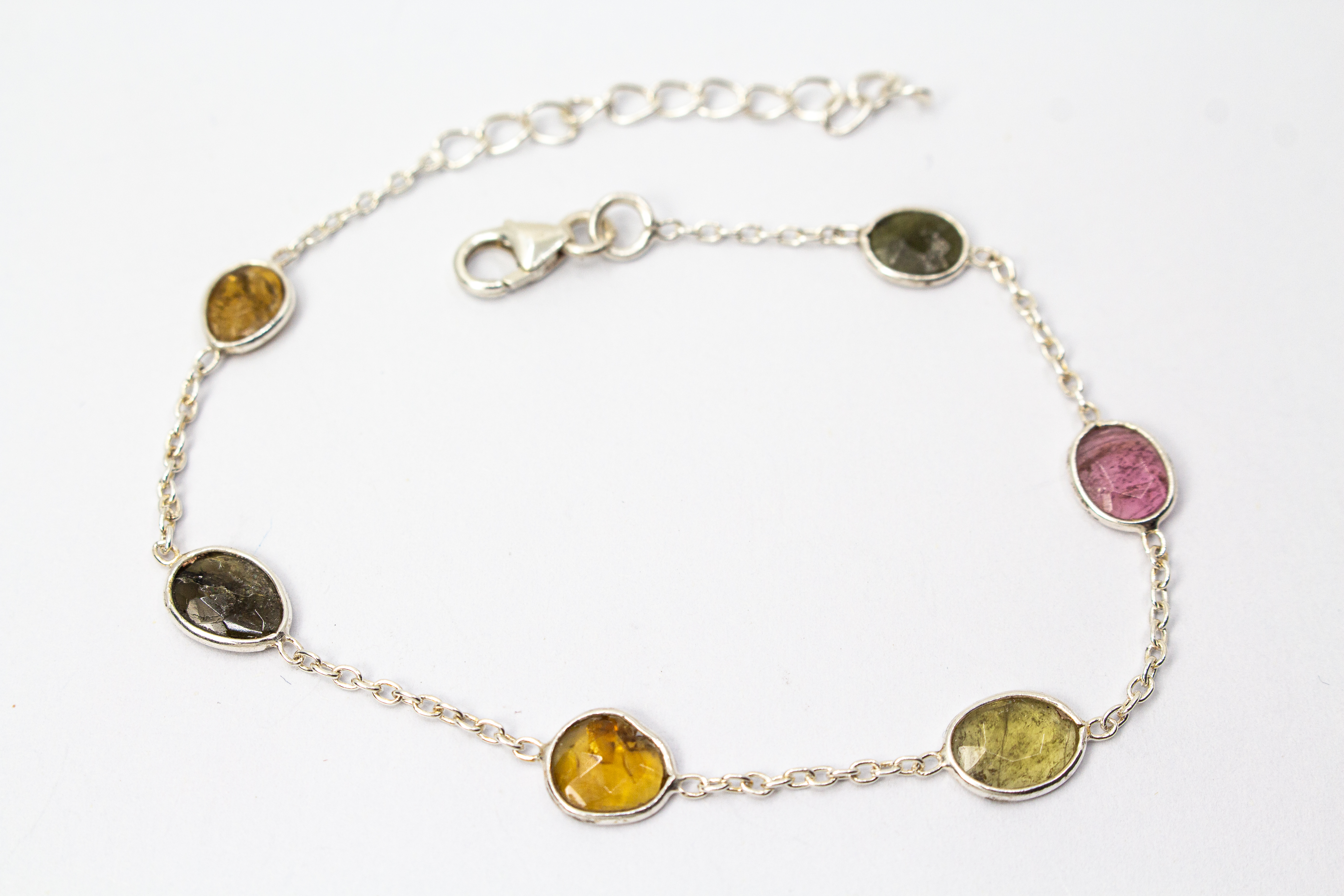 Ein filigranes Armband aus 925 Sterlingsilber von der Goldschmiede Regina Kraus, flach auf weißem Hintergrund arrangiert. In die feine Gliederkette sind sechs verschiedenfarbige, flache Turmaline in ovalen Zargenfassungen eingearbeitet – darunter Gelb-,