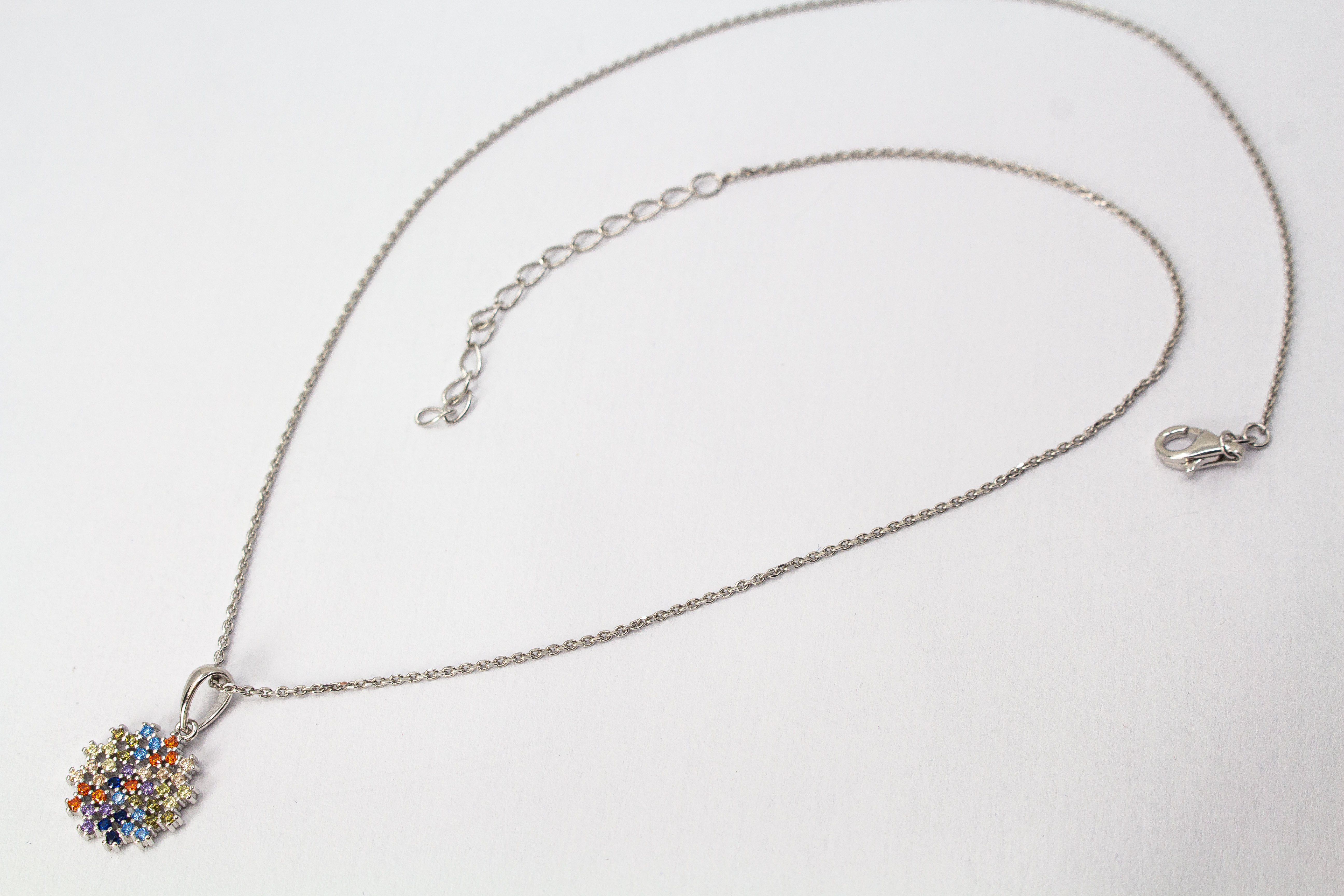Eine feine Halskette aus rhodiniertem 925er Sterlingsilber von der Goldschmiede Regina Kraus, schräg auf weißem Hintergrund liegend. Der runde Anhänger ist filigran wie ein Mandala oder eine Blüte gestaltet und dicht mit kleinen, bunt funkelnden Zirkonia-